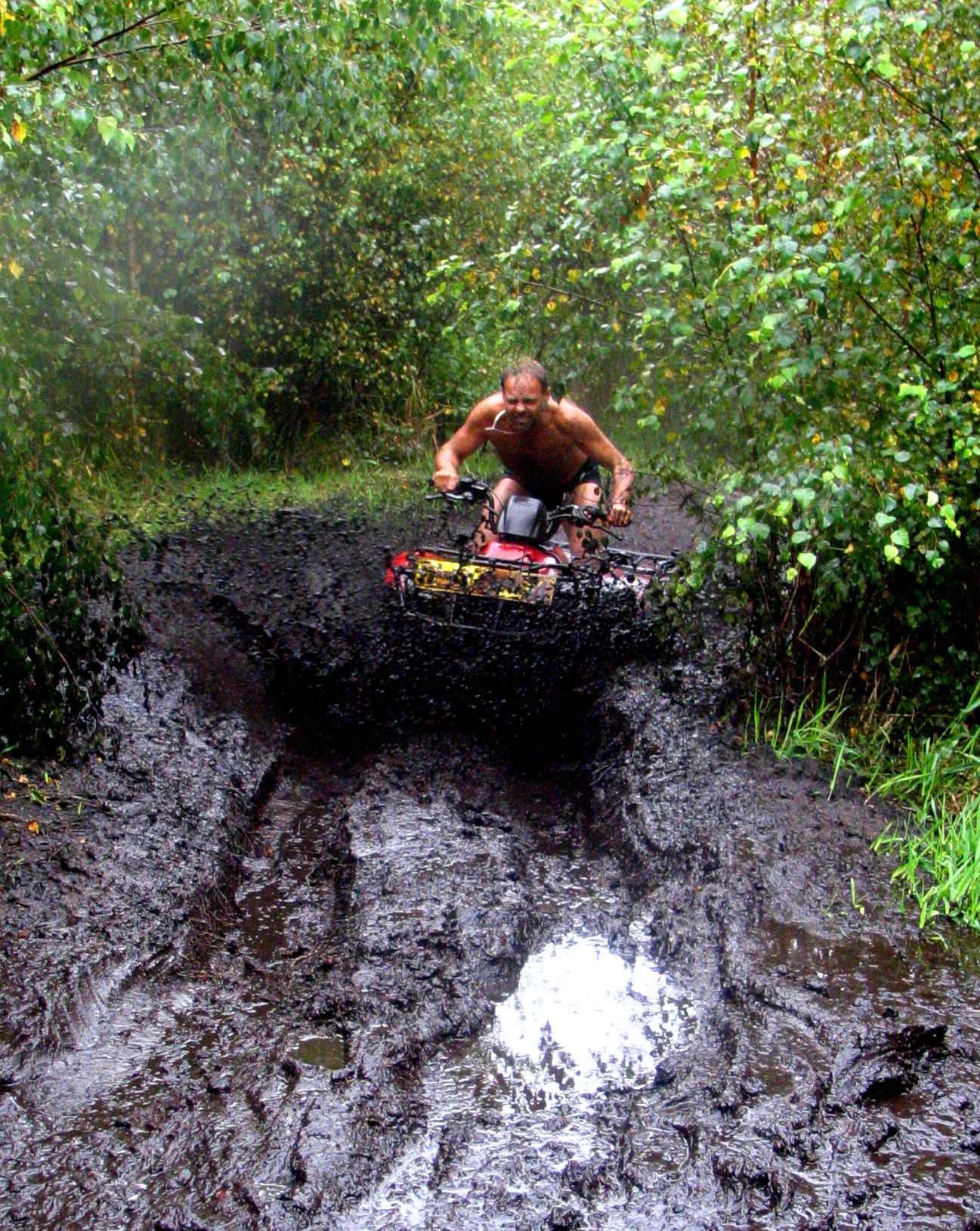 Mężczyzna bez koszulki, z papierosem w ustach, jadący czerwonym quadem przez błotnisty teren, w tle zielona roślinność.